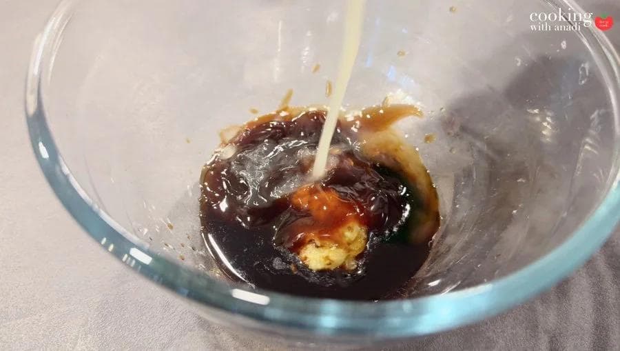 White liquid being poured into a glass bowl containing dark soy sauce, oyster sauce, and garlic paste mixture.