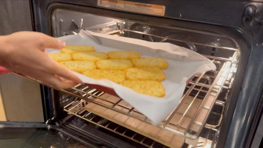Hand placing parchment paper with golden baked hash browns into an open oven.