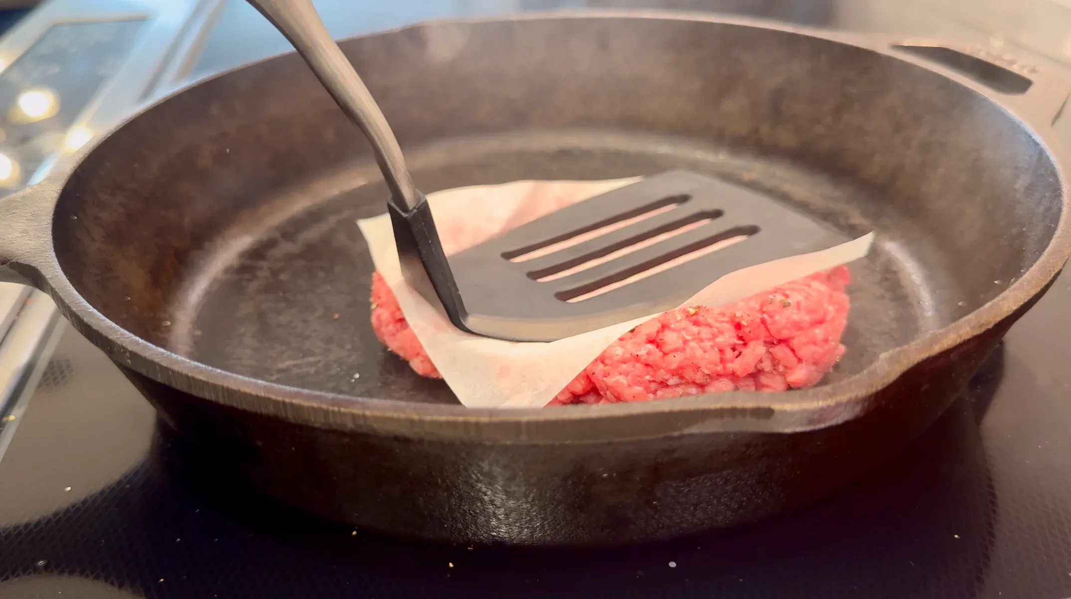 Ground beef patty being pressed down with a spatula in a cast iron pan lined with parchment paper.