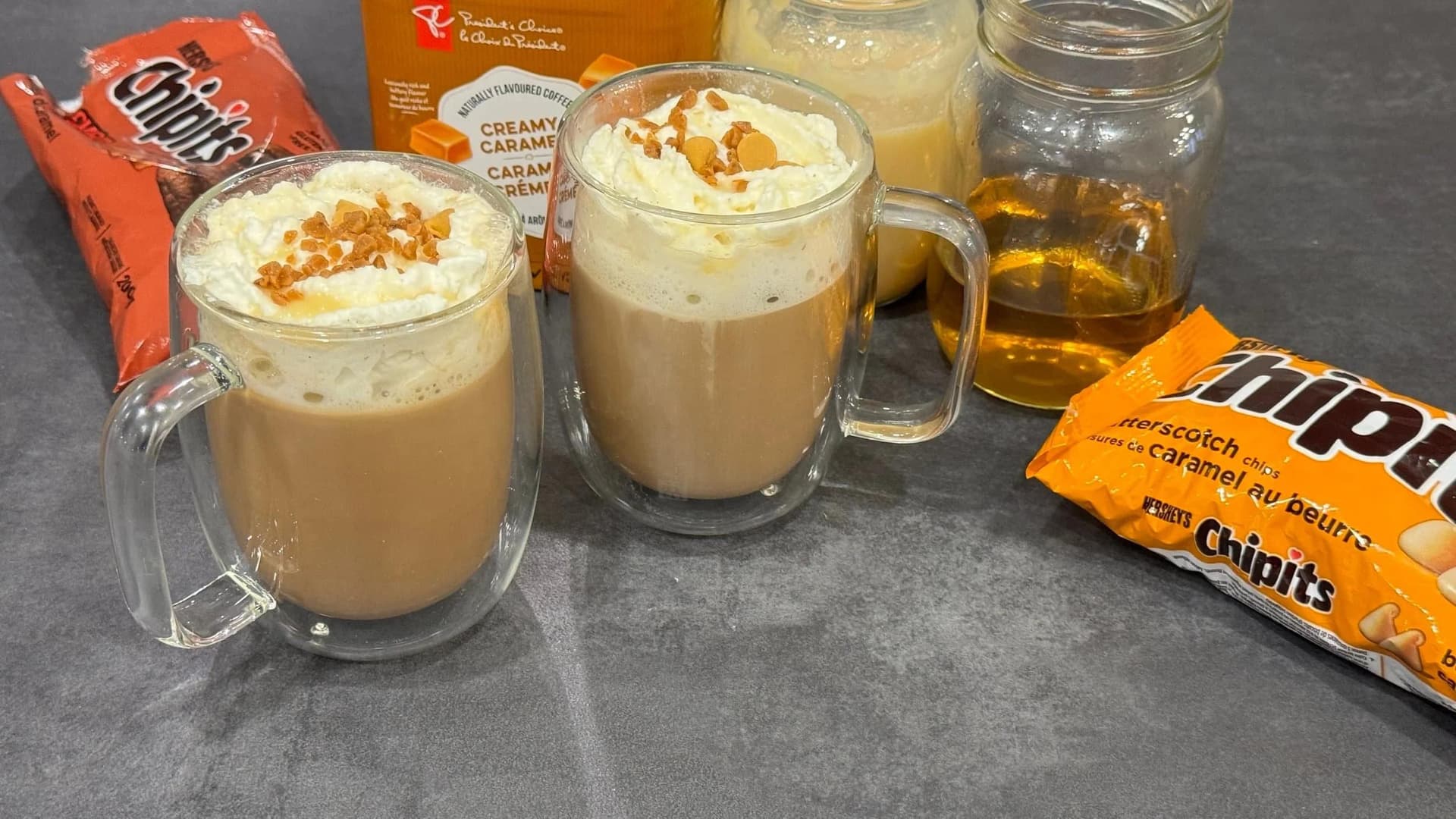 Two homemade caramel brulee lattes in glass mugs topped with whipped cream and caramel bits, surrounded by ingredients.