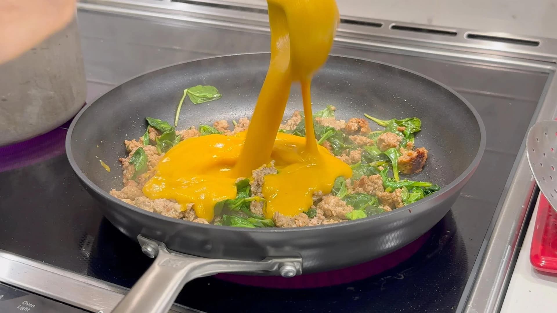 Butternut squash sauce being poured into a pan with cooked ground sausage and spinach on a stovetop.