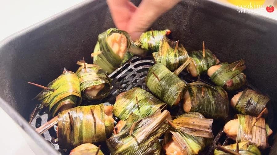 Pandan-wrapped chicken pieces secured with toothpicks arranged in an air fryer basket, being placed by hand.