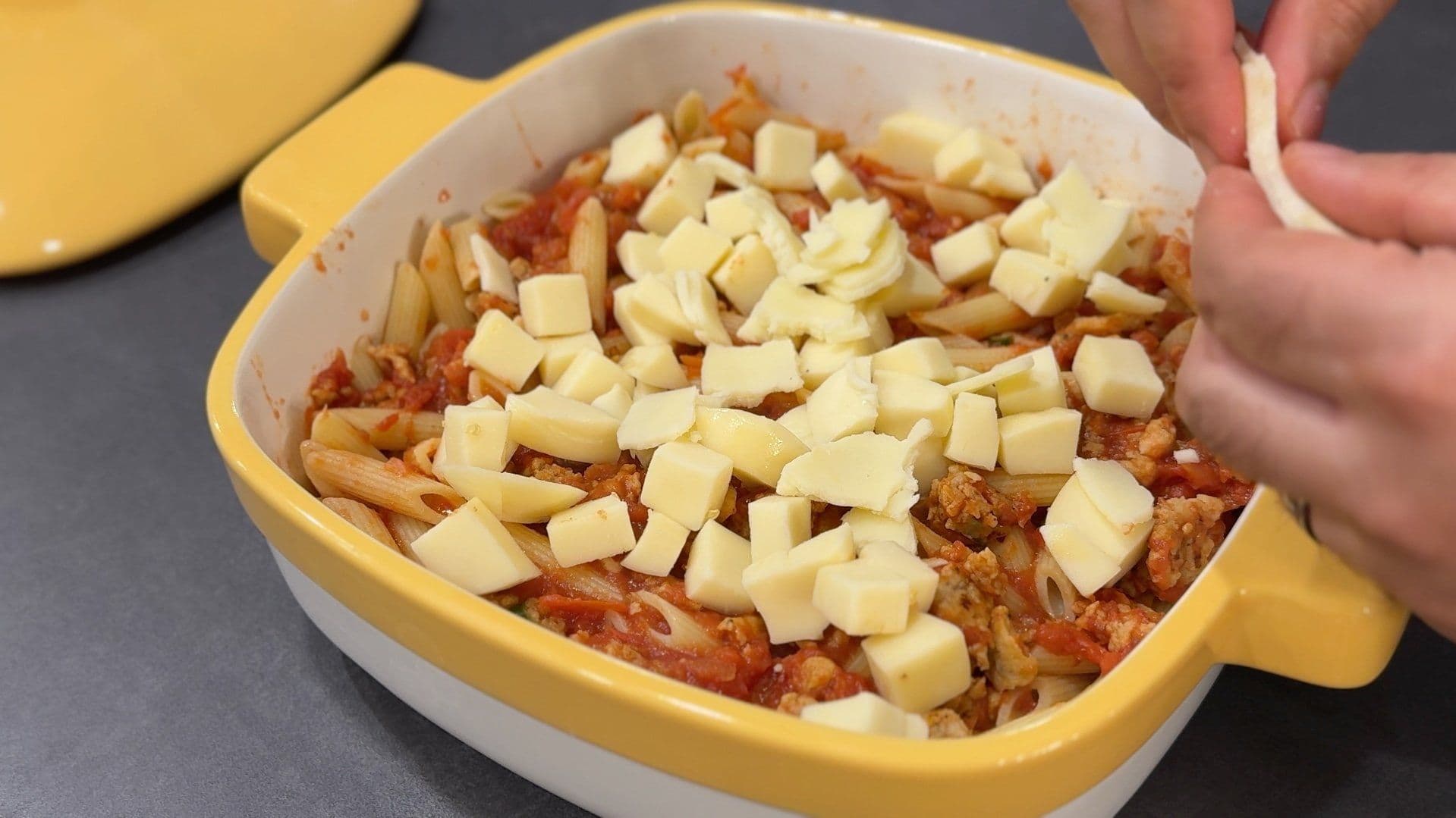 Baked penne casserole topped with cubed mozzarella cheese before baking in a yellow baking dish.