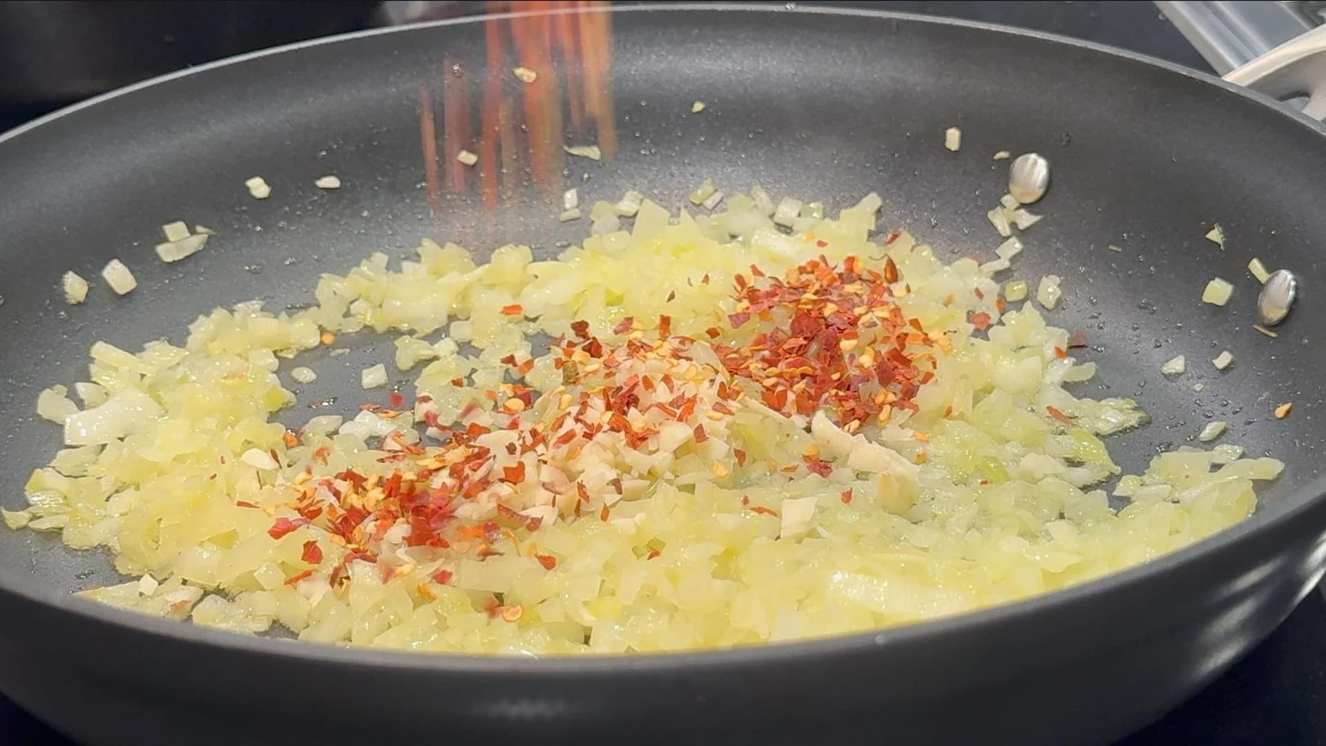 Diced onions sautéing in a dark skillet with red chilli flakes just added in the center.