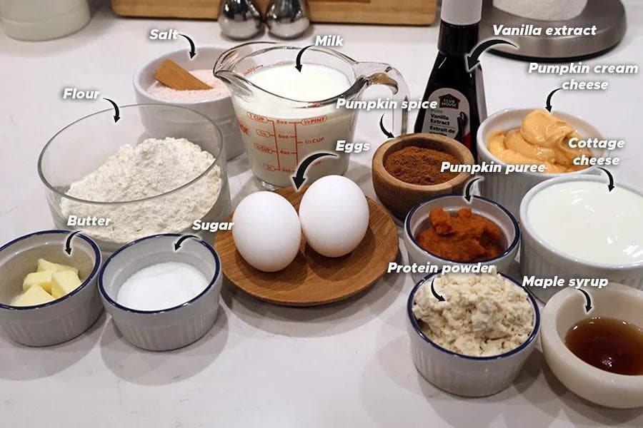 Ingredients for high protein pumpkin crepes laid out in bowls and containers on a white surface, including flour, eggs, pumpkin puree, and spices.