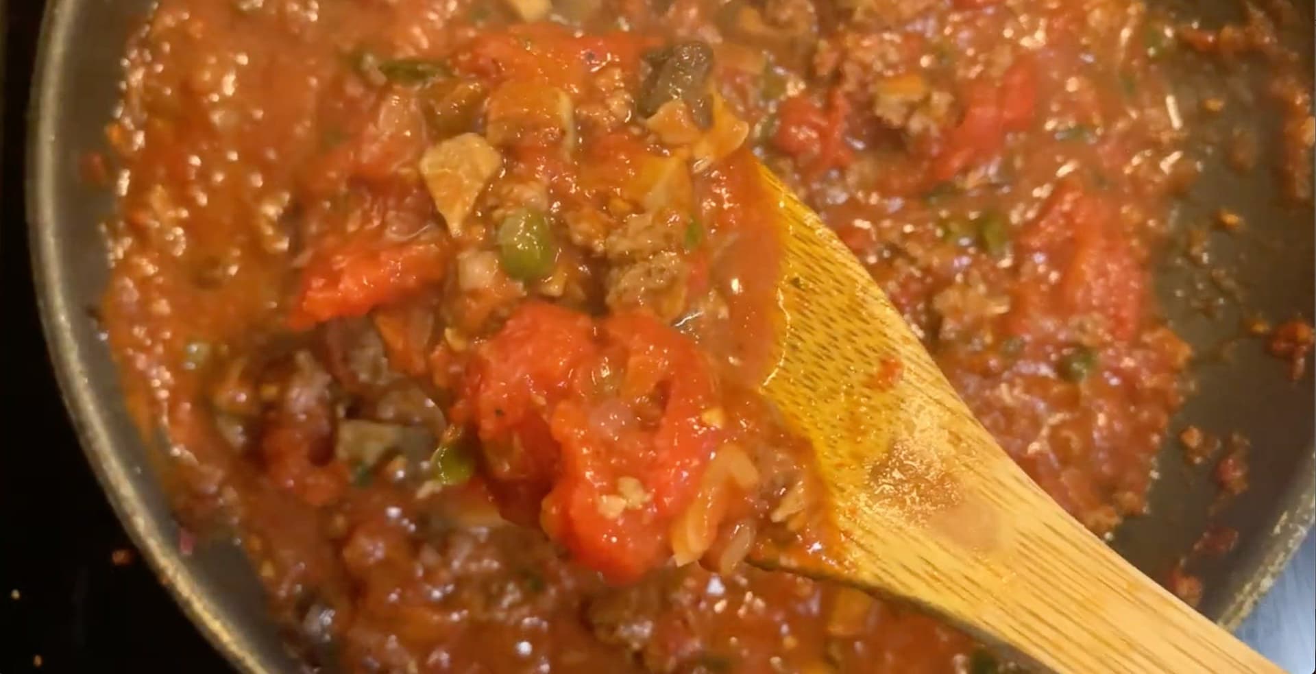 Meat sauce simmering in a pot with tomatoes, ground beef, and green peppers being stirred with a wooden spoon.