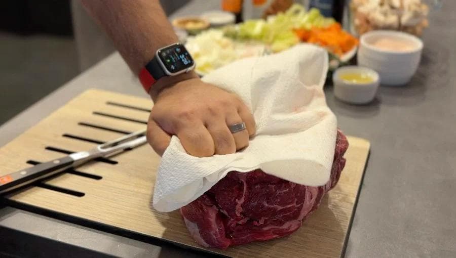 Hand patting dry a raw beef roast with paper towels on a wooden cutting board before searing.