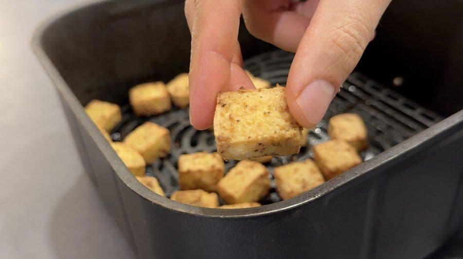 Hand placing a golden-brown tofu cube into an air fryer basket filled with crispy tofu pieces.