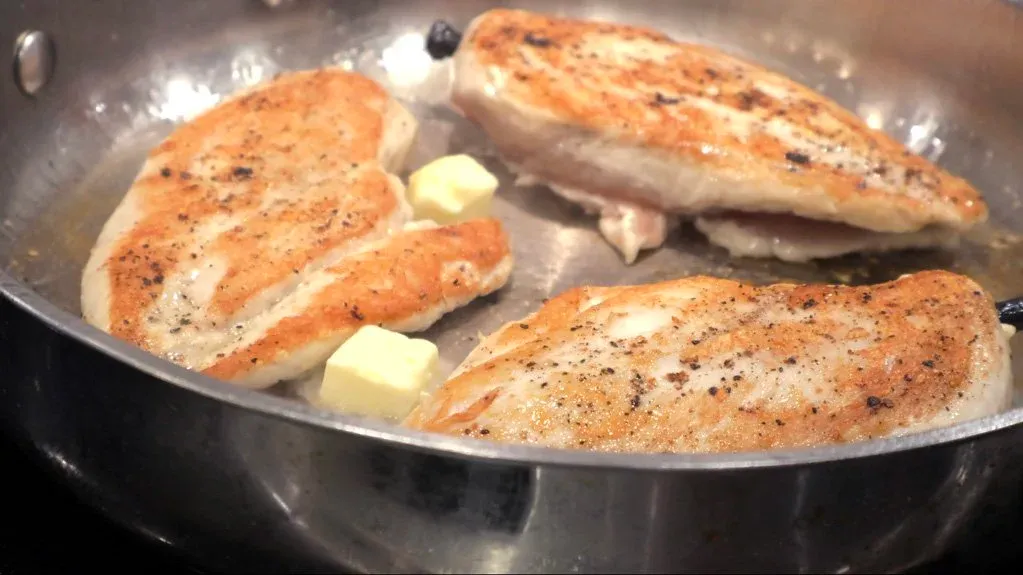 Three seasoned chicken breasts searing in a pan with butter and garlic cloves.