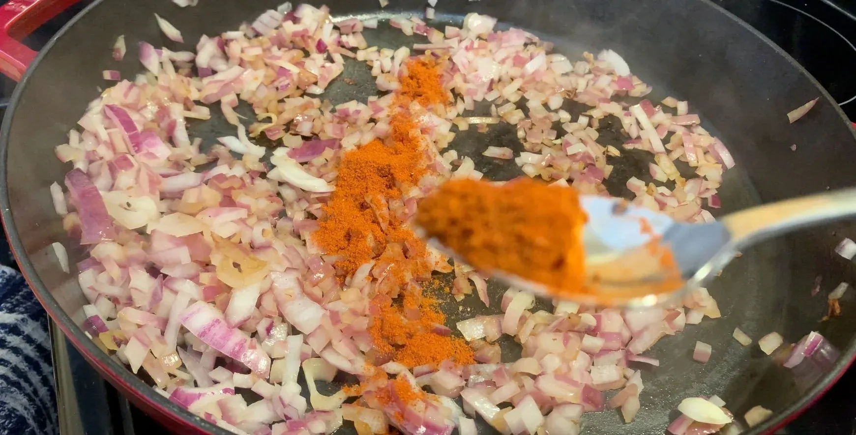 Diced onions and spices including paprika and cayenne being added to a pan with a wooden spoon.