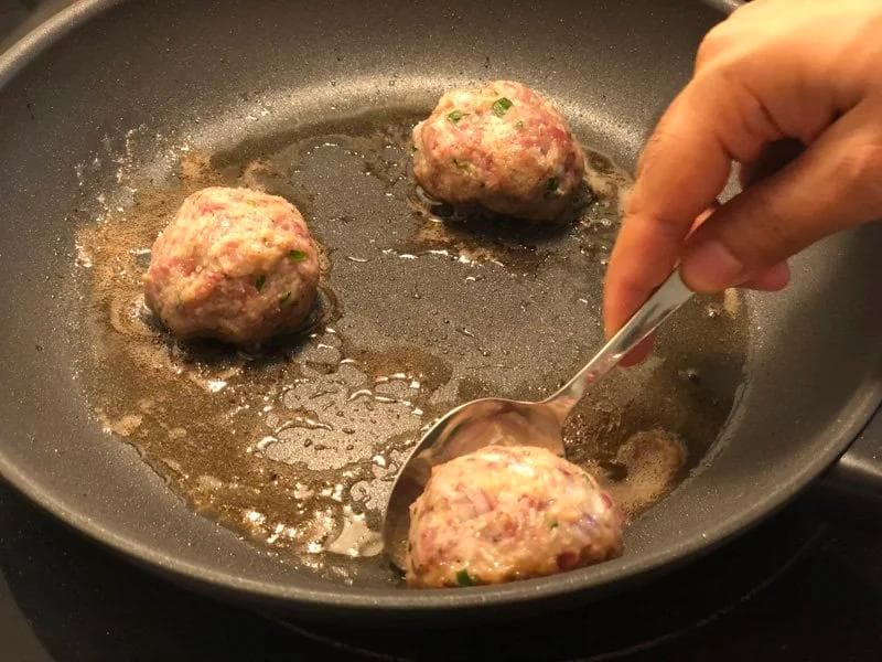 Three meatballs browning in a skillet with oil, one being turned with a spoon.