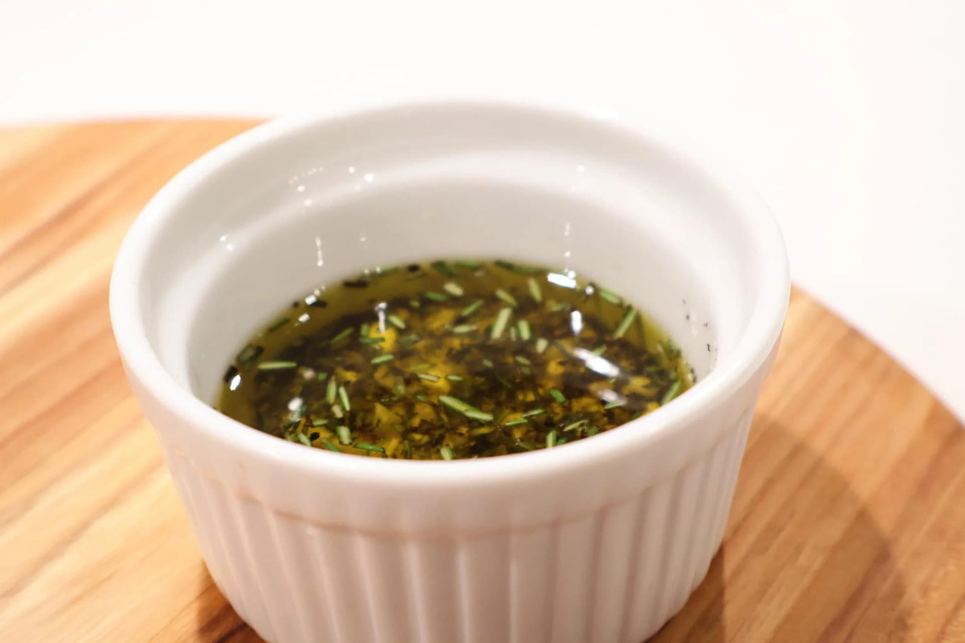 White ramekin filled with olive oil infused with fresh rosemary and mixed herbs, resting on a wooden board.