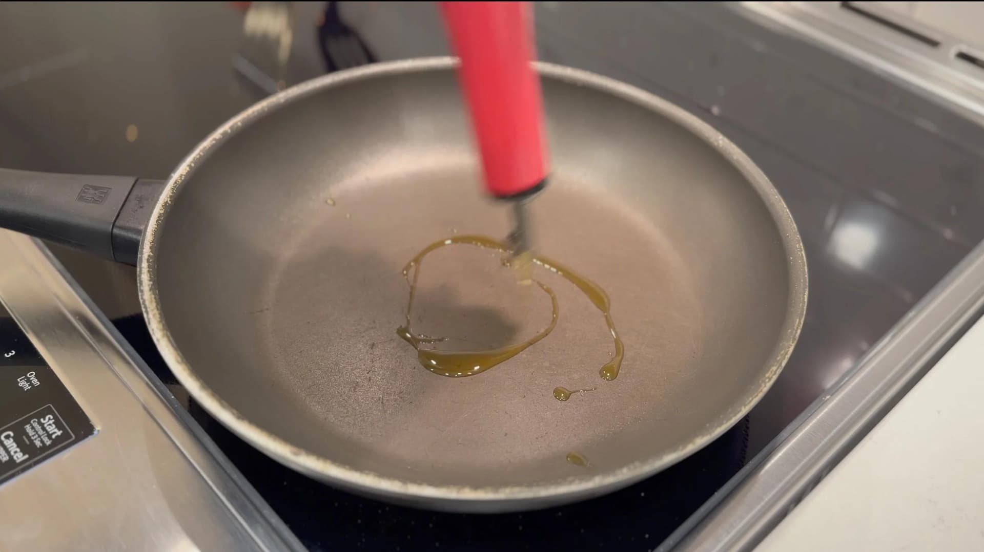 Olive oil heating in a pan on a stovetop, forming a small pool in the center of the pan.