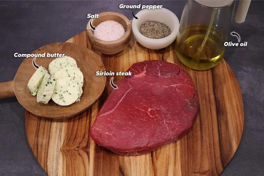 Ingredients for air fryer steak displayed on a wooden board: sirloin steak, compound butter, salt, ground pepper, and olive oil.