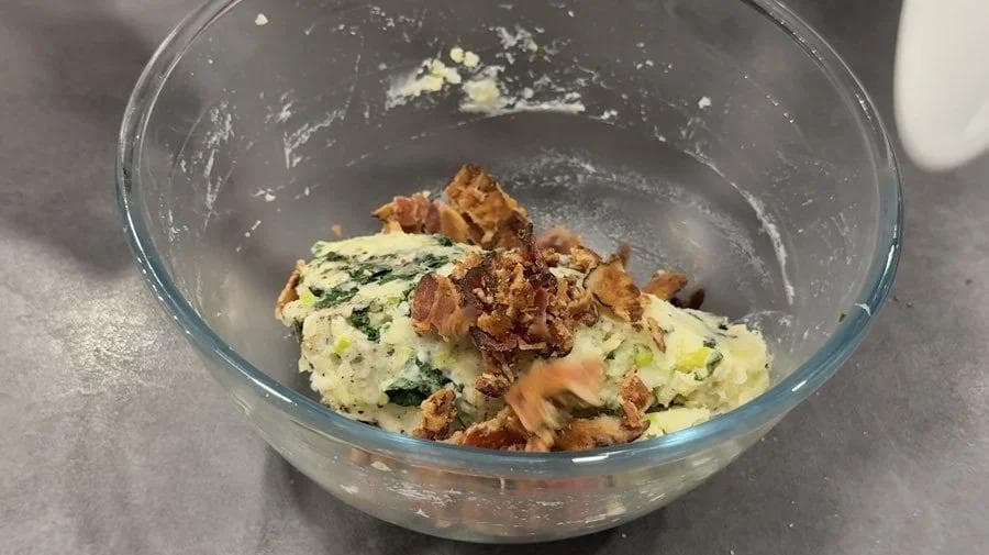 Colcannon filling with bacon bits mixed into creamy mashed potatoes with herbs in a glass bowl.