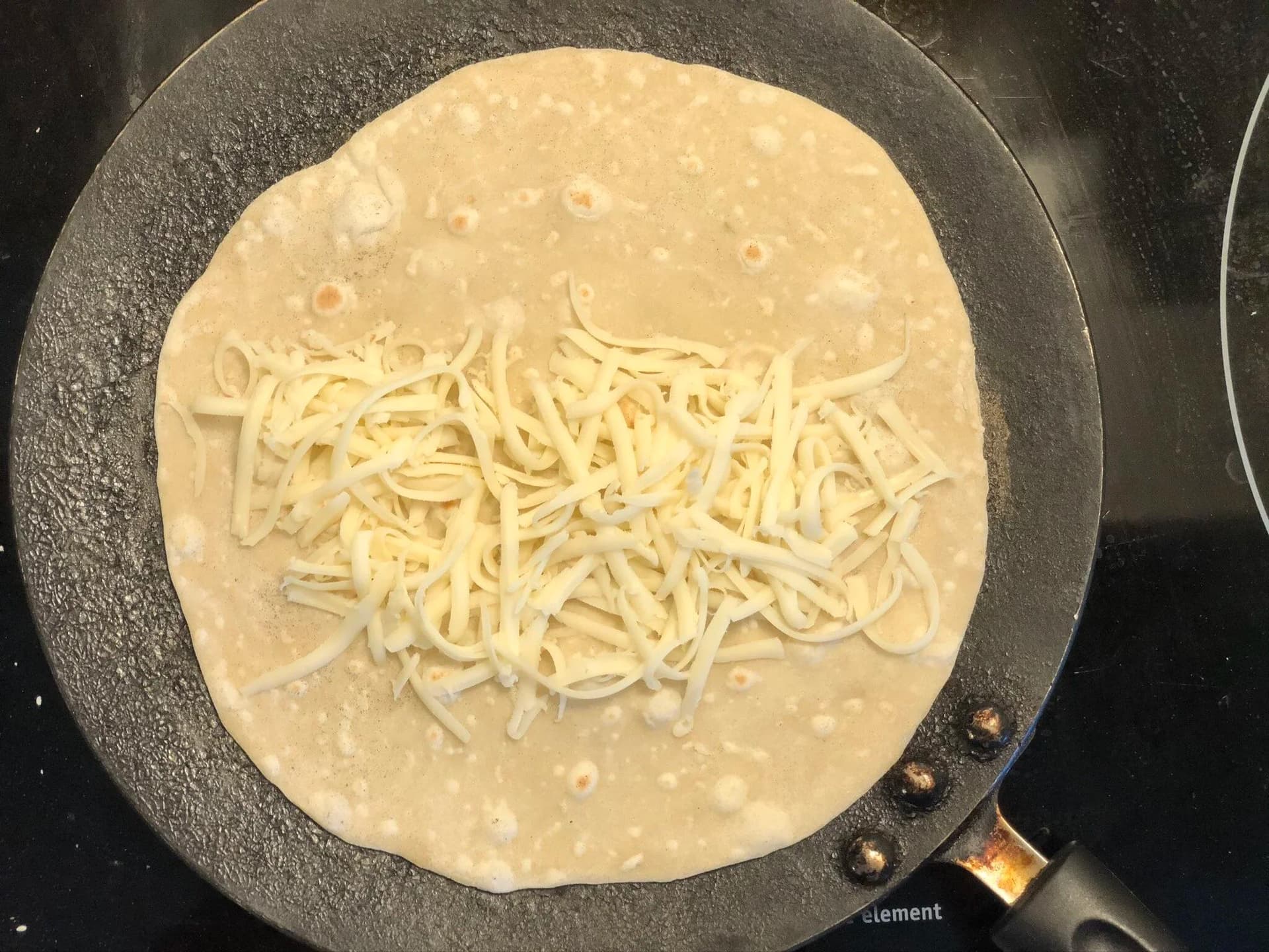 Shredded cheese melting in center of tortilla cooking on dark skillet.
