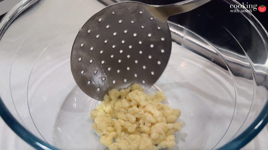 Cooked spätzle being removed from boiling water with a slotted spoon held over a glass bowl.