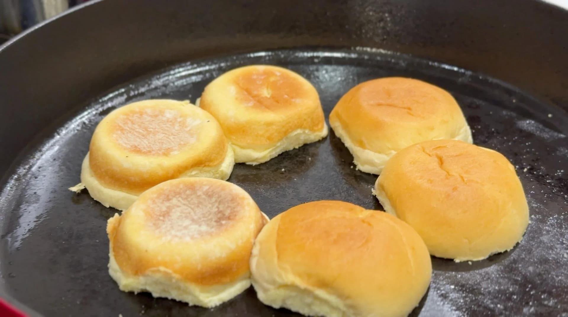 Six toasted burger buns with golden-brown tops in a cast iron skillet with oil.