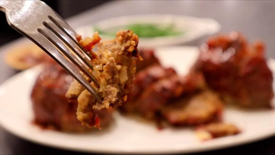 Fork lifting a golden-brown chipotle mini meatloaf with red peppers from a white plate with more meatloaves blurred in background.