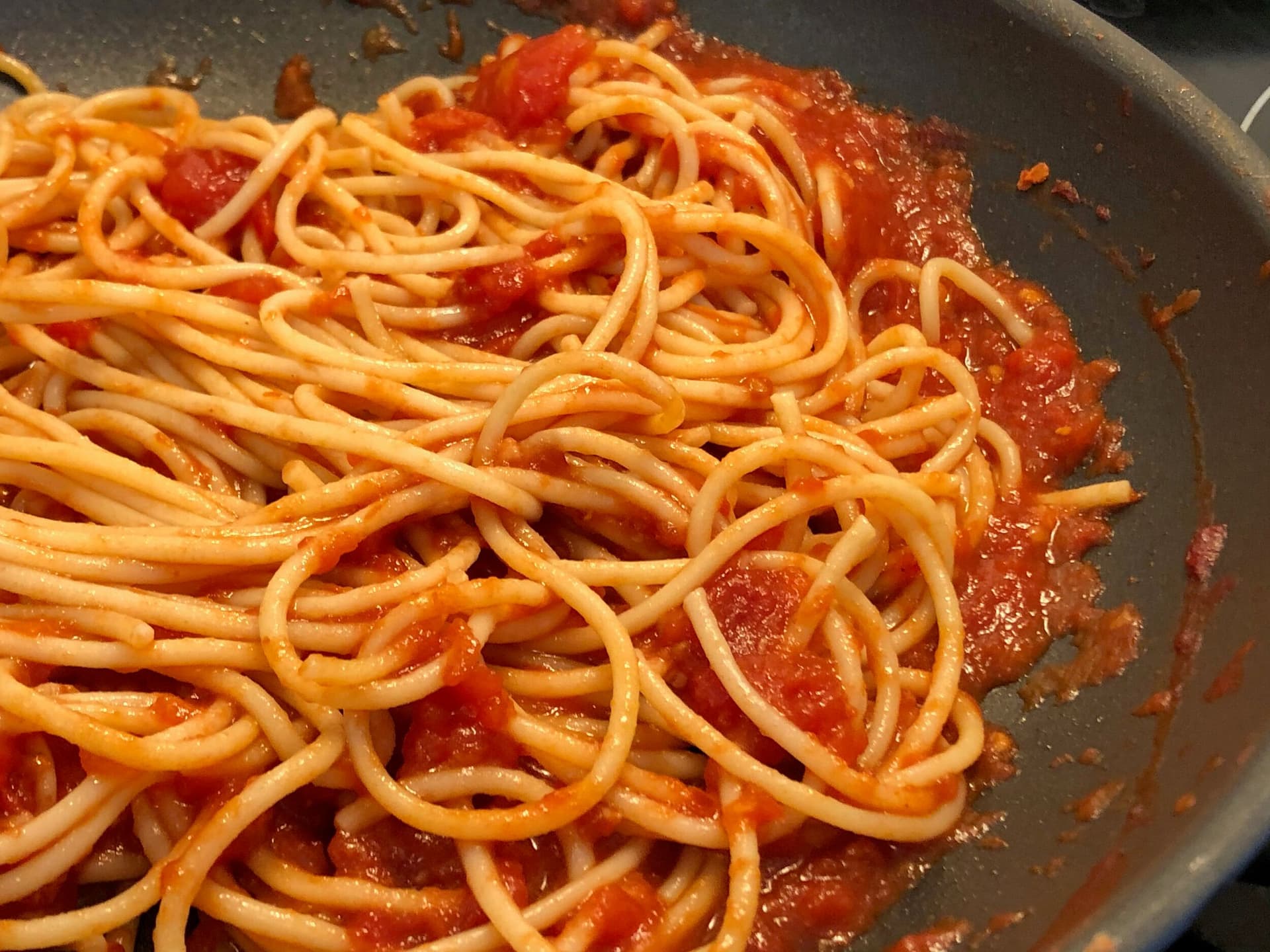 Spaghetti coated in marinara sauce being tossed together in a dark pan.