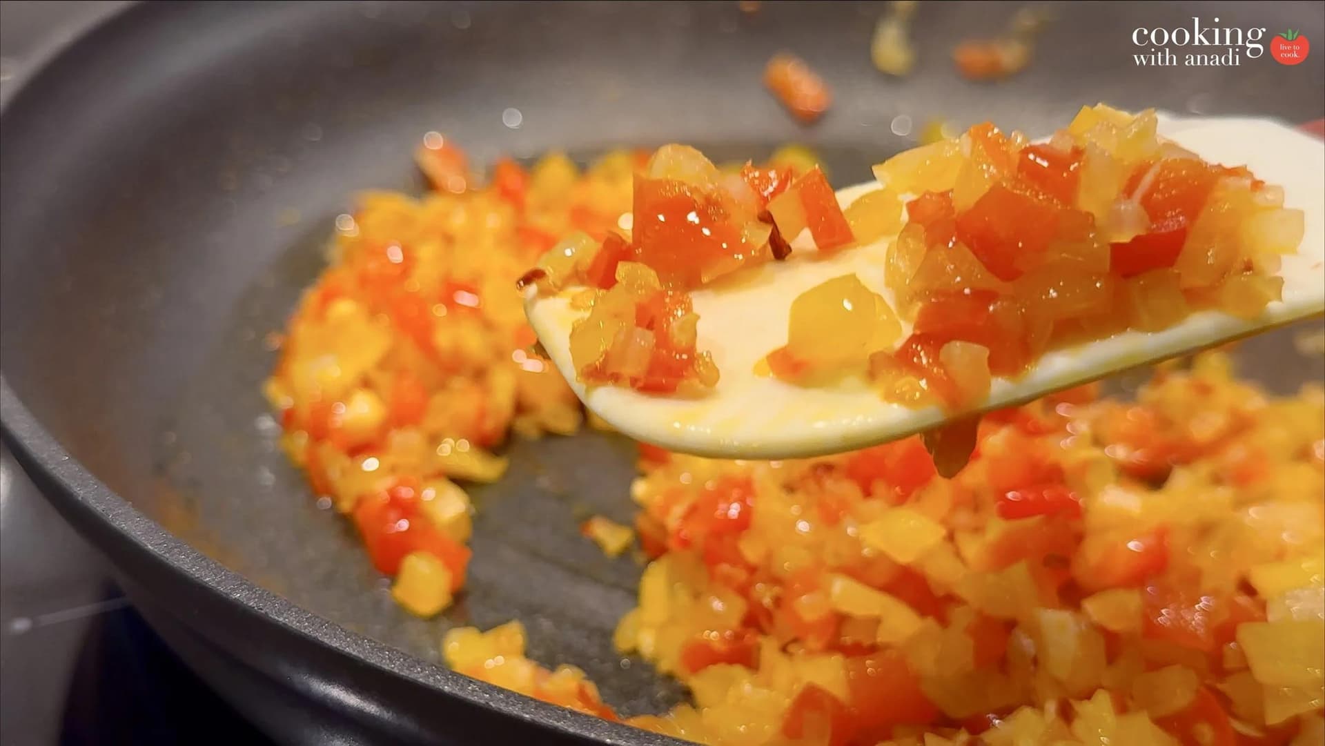 Diced red and yellow bell peppers sautéed until softened in a dark skillet, shown on a wooden spoon.