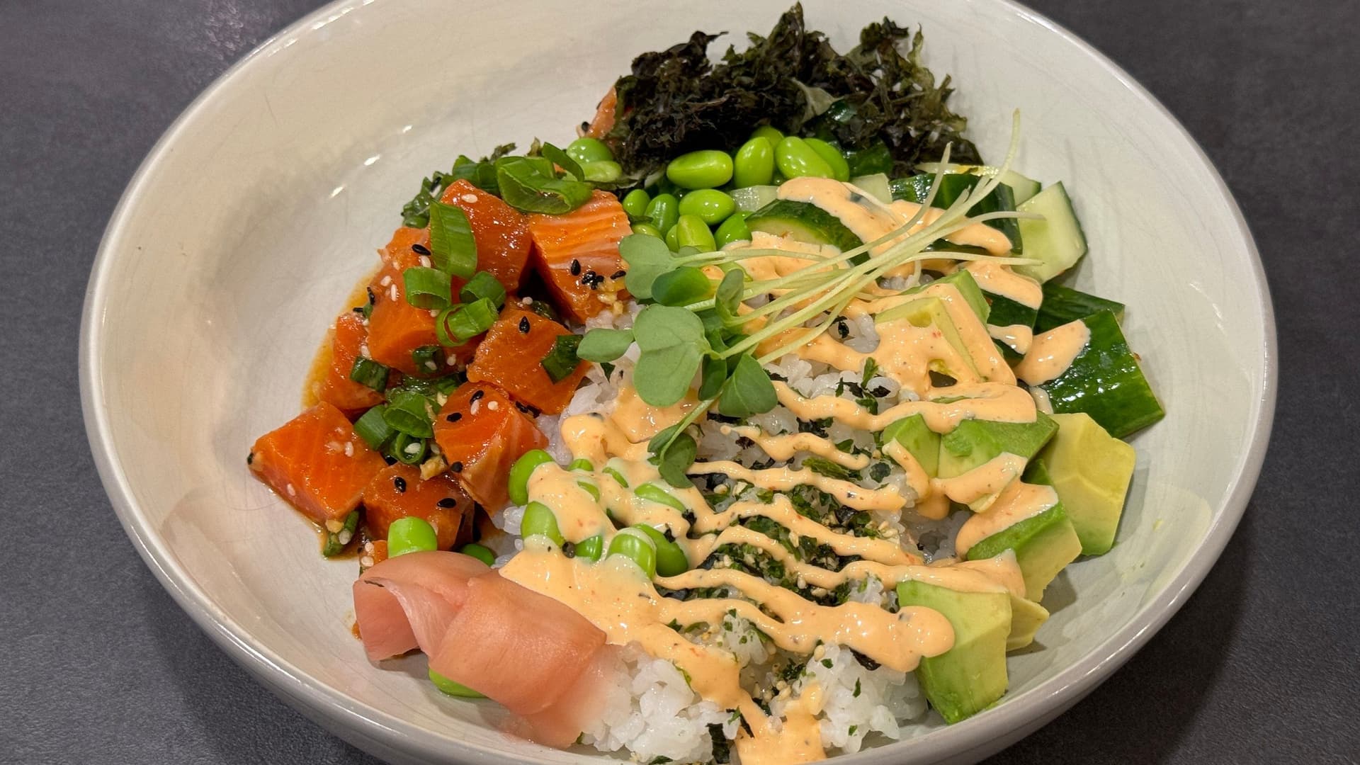Salmon poke bowl with rice, avocado, cucumber, red pepper, sesame seeds, seaweed, and spicy mayo drizzle in white bowl.