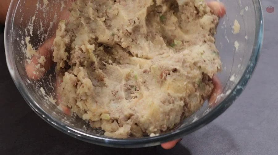 Meat and potato mixture being mixed in a glass bowl with hands visible, showing a crumbly, combined texture ready for cooling.