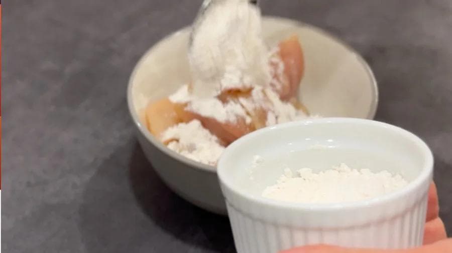 Chicken pieces being coated with flour in a white bowl for country harvest stew.