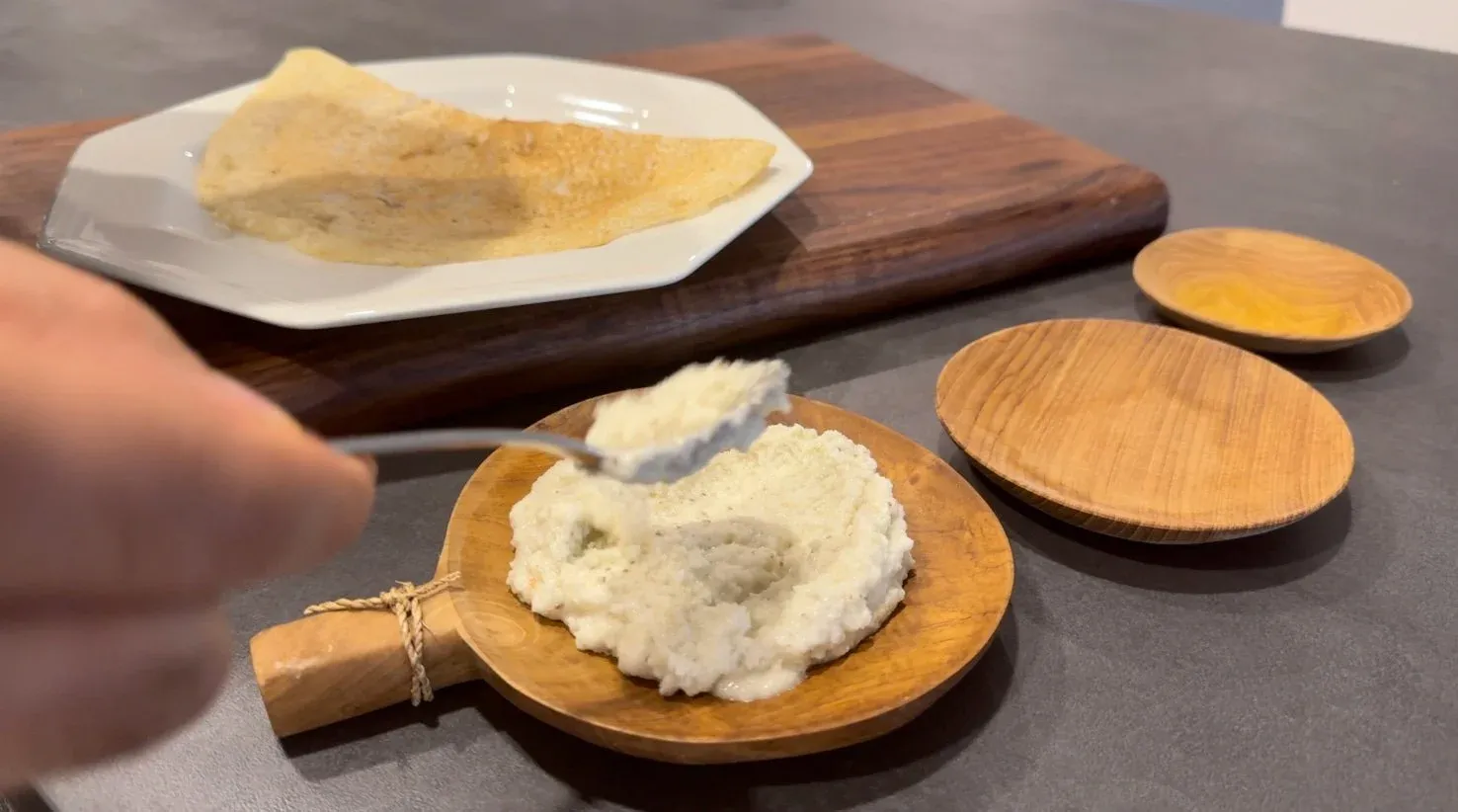 Coconut chutney in wooden serving spoon with crepe on plate and wooden utensils nearby.