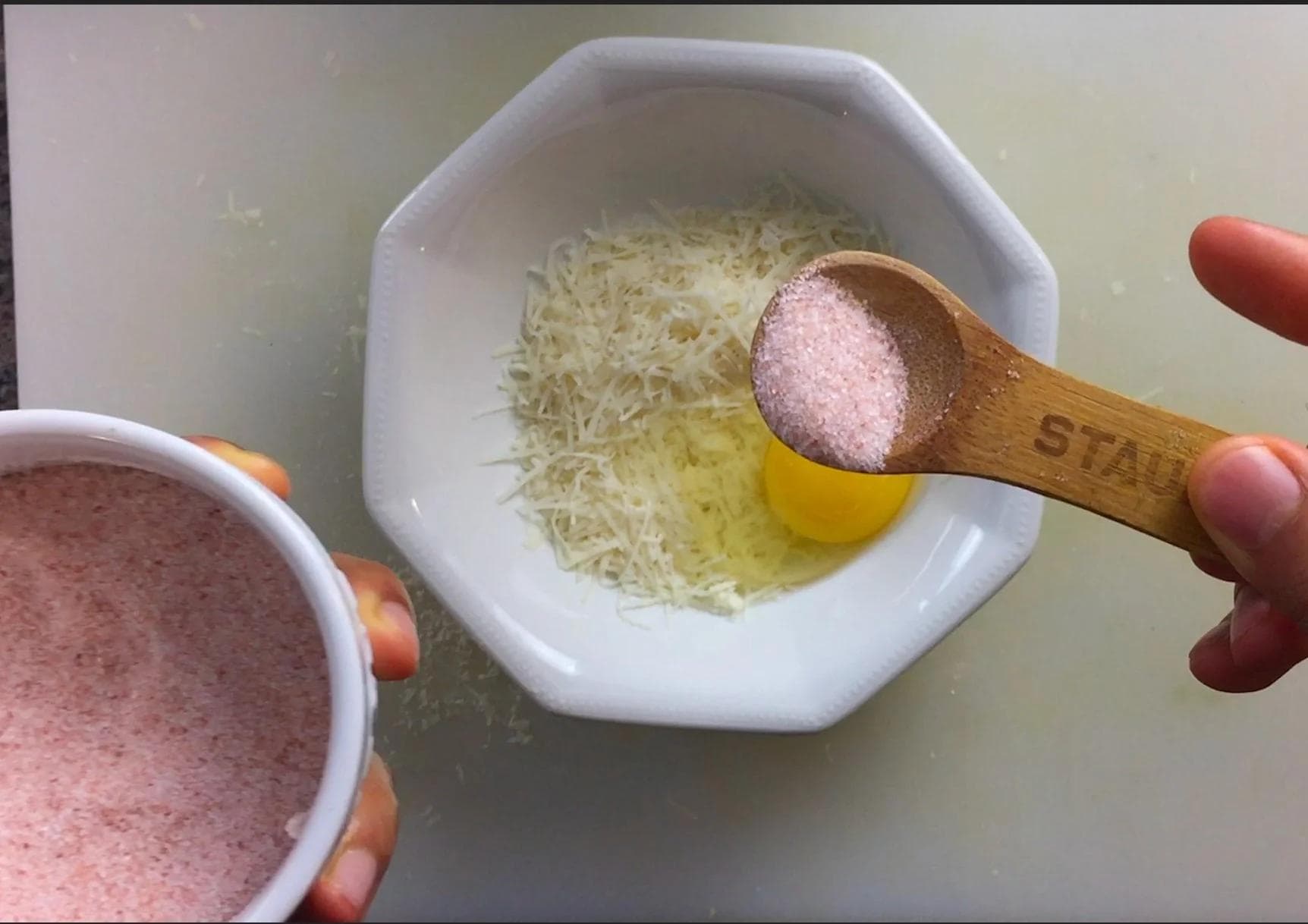 Wooden spoon holding pink salt over a white bowl with shredded parmesan cheese and egg yolk, with a bowl of egg mixture visible on the left.