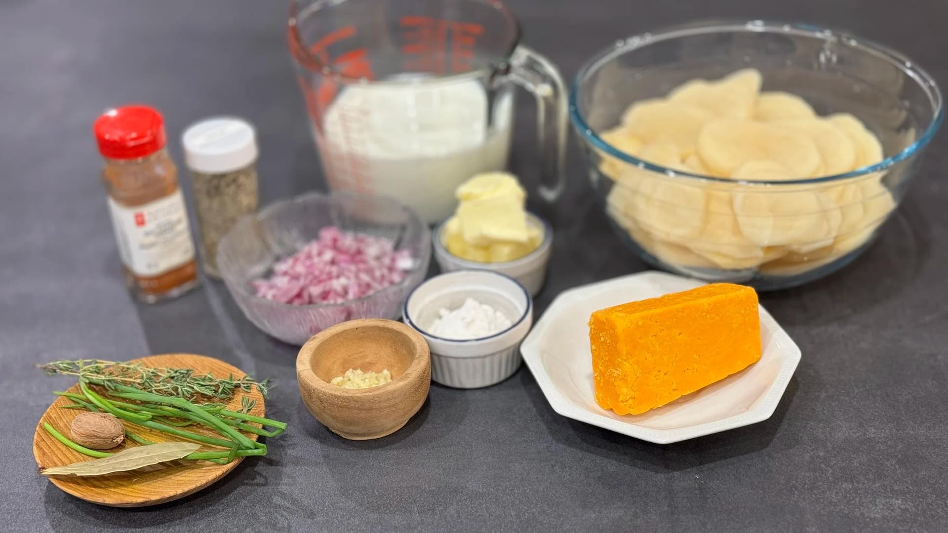 Ingredients for scalloped potatoes including sliced potatoes, cheddar cheese, milk, butter, garlic, thyme, and seasonings arranged on a dark surface.