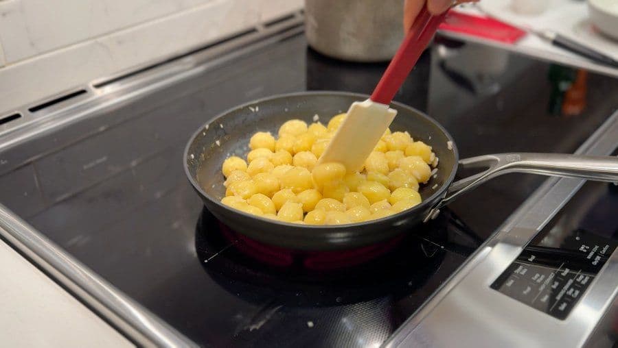 Butter-coated baby potatoes being stirred in a pan on an electric stovetop with a wooden spatula.