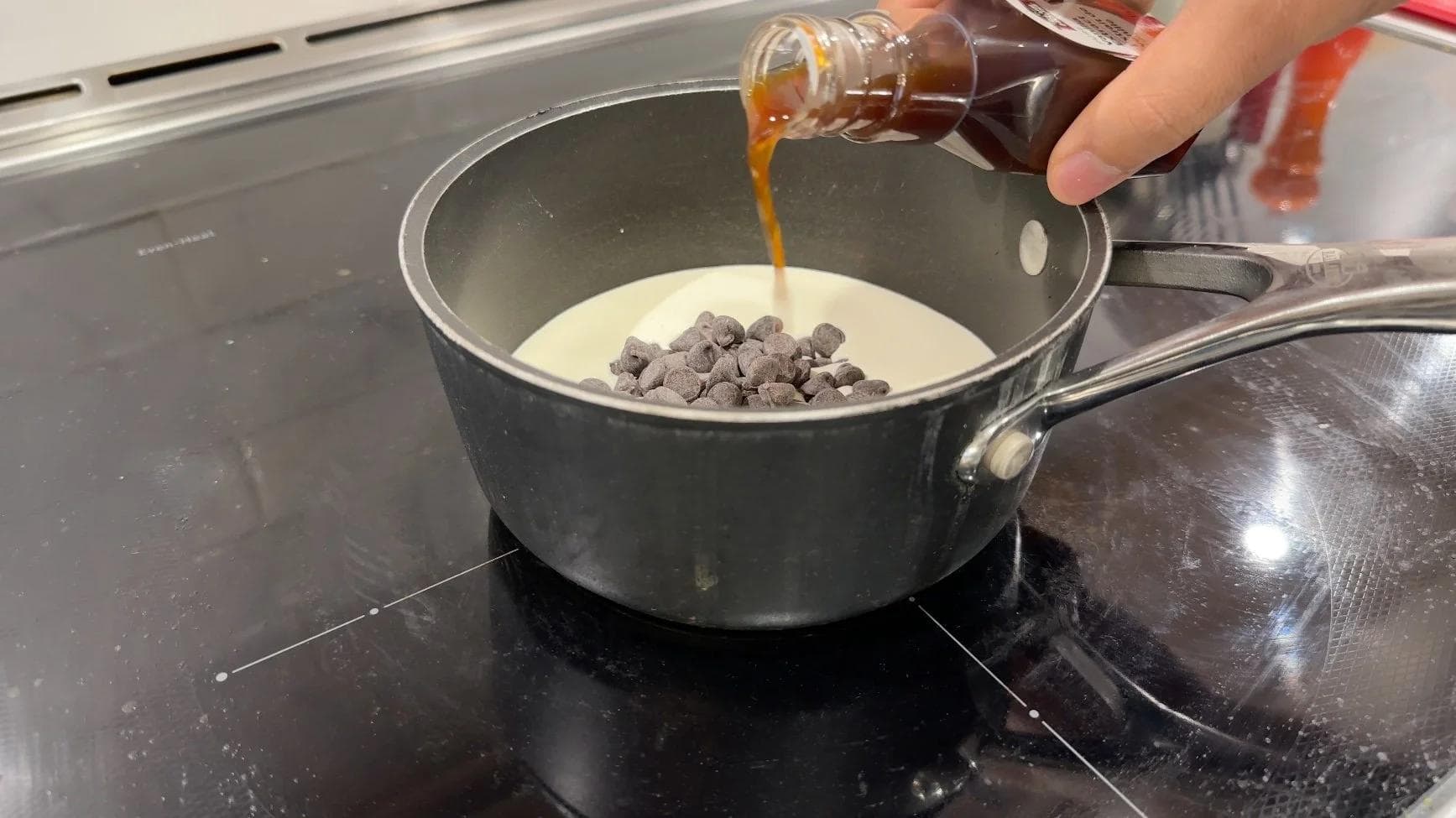 Pouring caramel sauce into a saucepan with cream and chocolate chips on a stovetop.