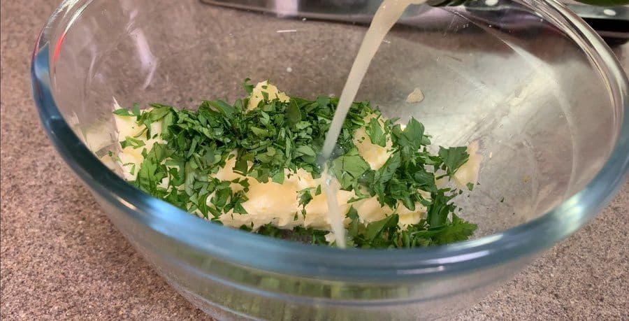 Chopped fresh parsley and minced garlic being stirred into pale butter in a glass bowl with a spatula.