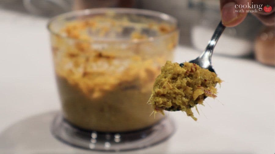 Spice paste being lifted on a spoon from a food processor, showing thick, golden-yellow curry paste texture.