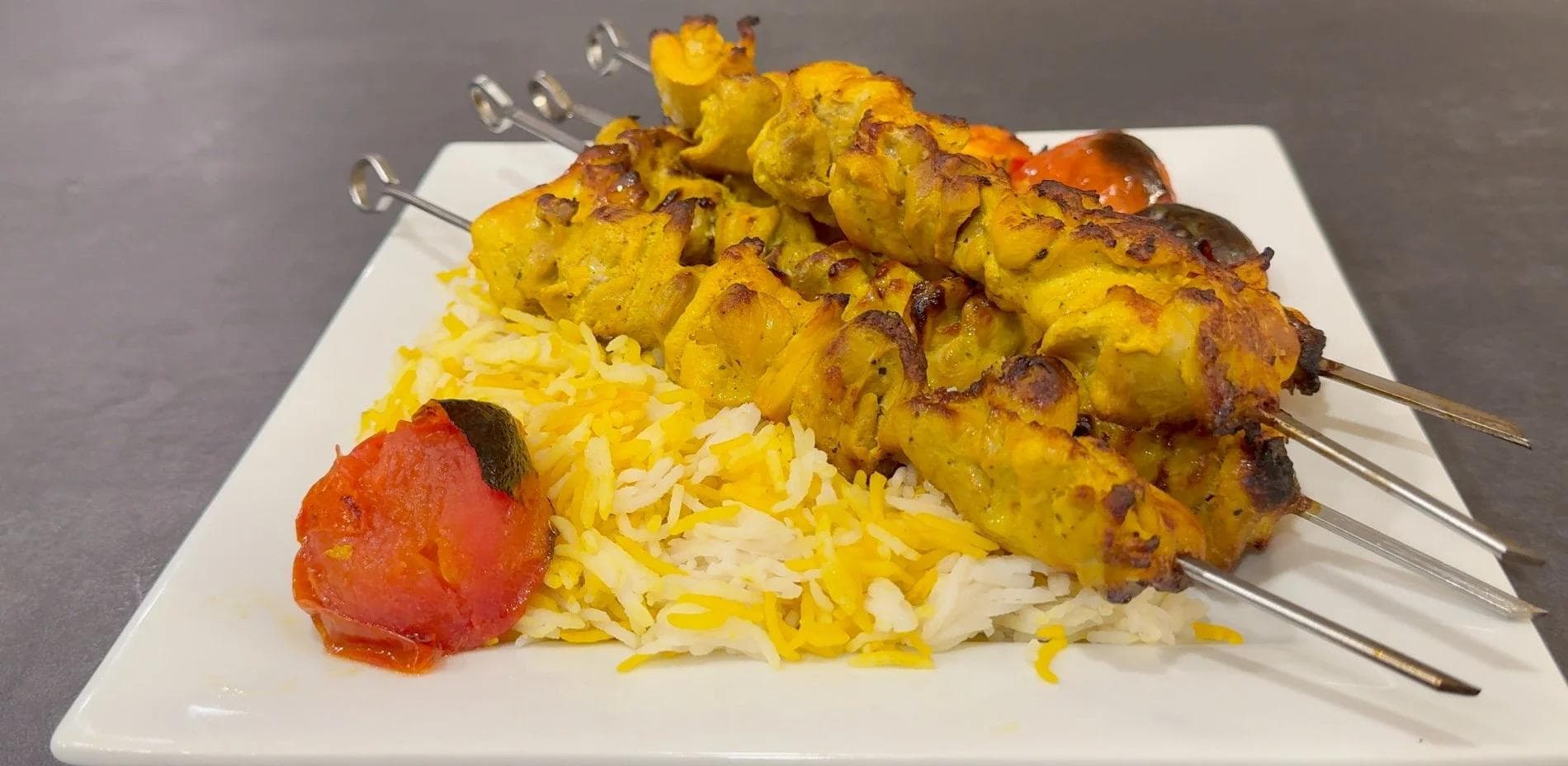 Joojeh kebab skewers served over saffron rice with roasted tomato on white plate.