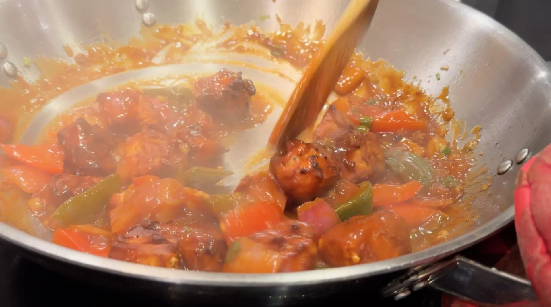 Chilli paneer tossing in thick tomato sauce with bell peppers in a stainless steel wok, stirred with a wooden spoon.