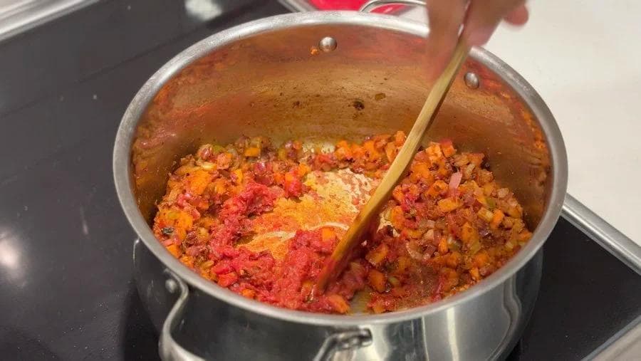 Tomato paste being stirred into a pot of diced vegetables and spices with a wooden spoon.