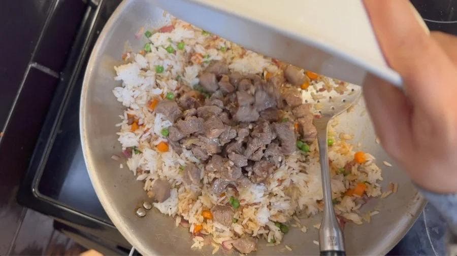 Beef fried rice in a white bowl with rice, ground beef, diced vegetables, and green onions, being stirred with a spoon.