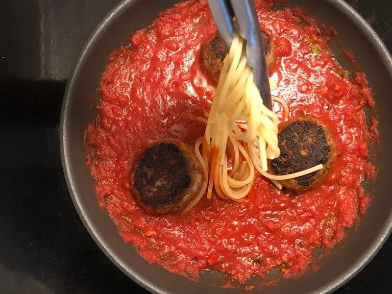 Spaghetti being added to a pan of marinara sauce with cooked meatballs simmering together.