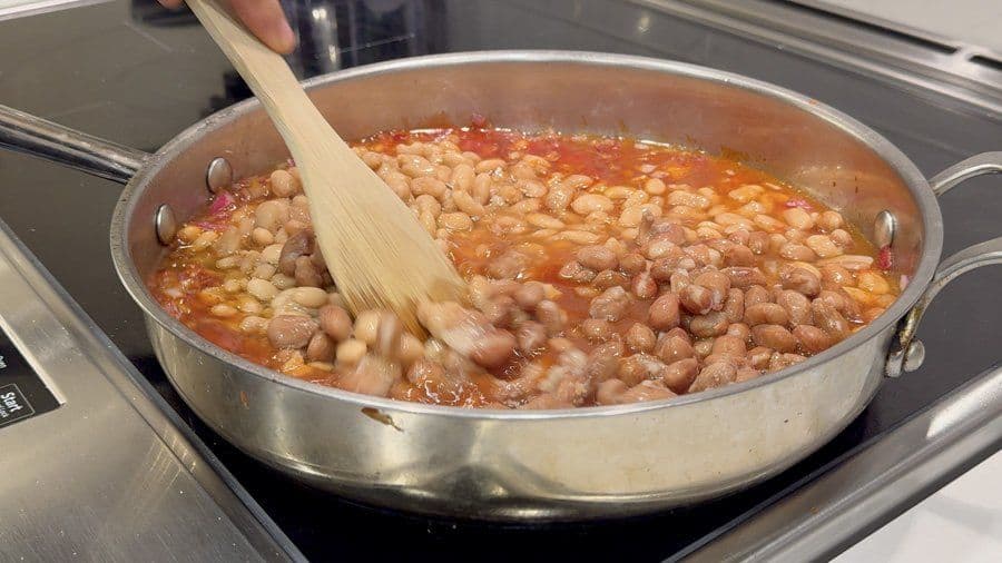 Wooden spoon stirring a pot of baked beans with pinto and great northern beans in a rich tomato-based sauce.