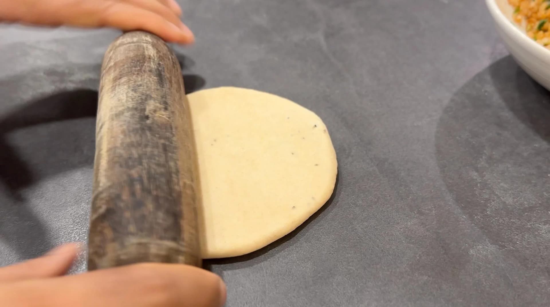 Rolling dough disk for gobi paratha with wooden rolling pin on dark surface.