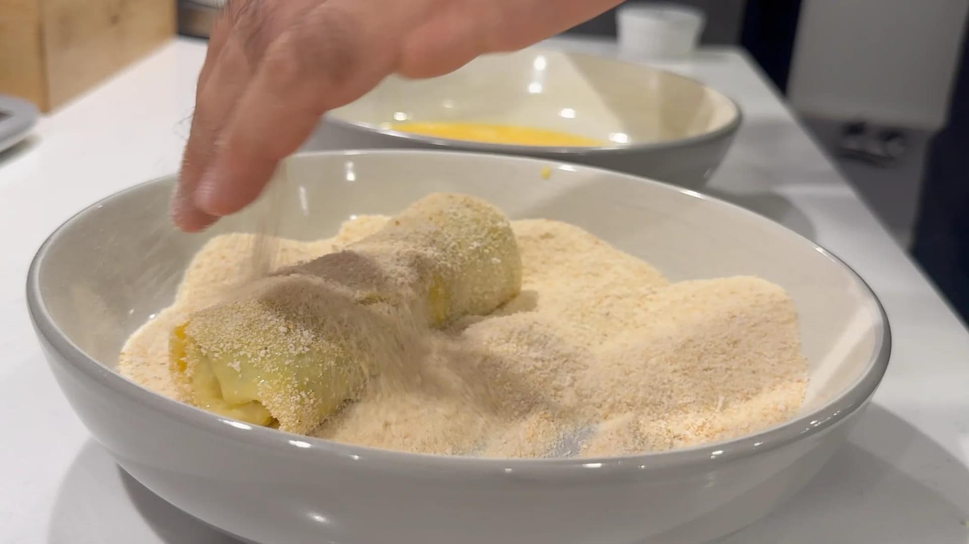 Breaded krokiety rolls being coated in breadcrumbs at a breading station with egg wash bowl in background.