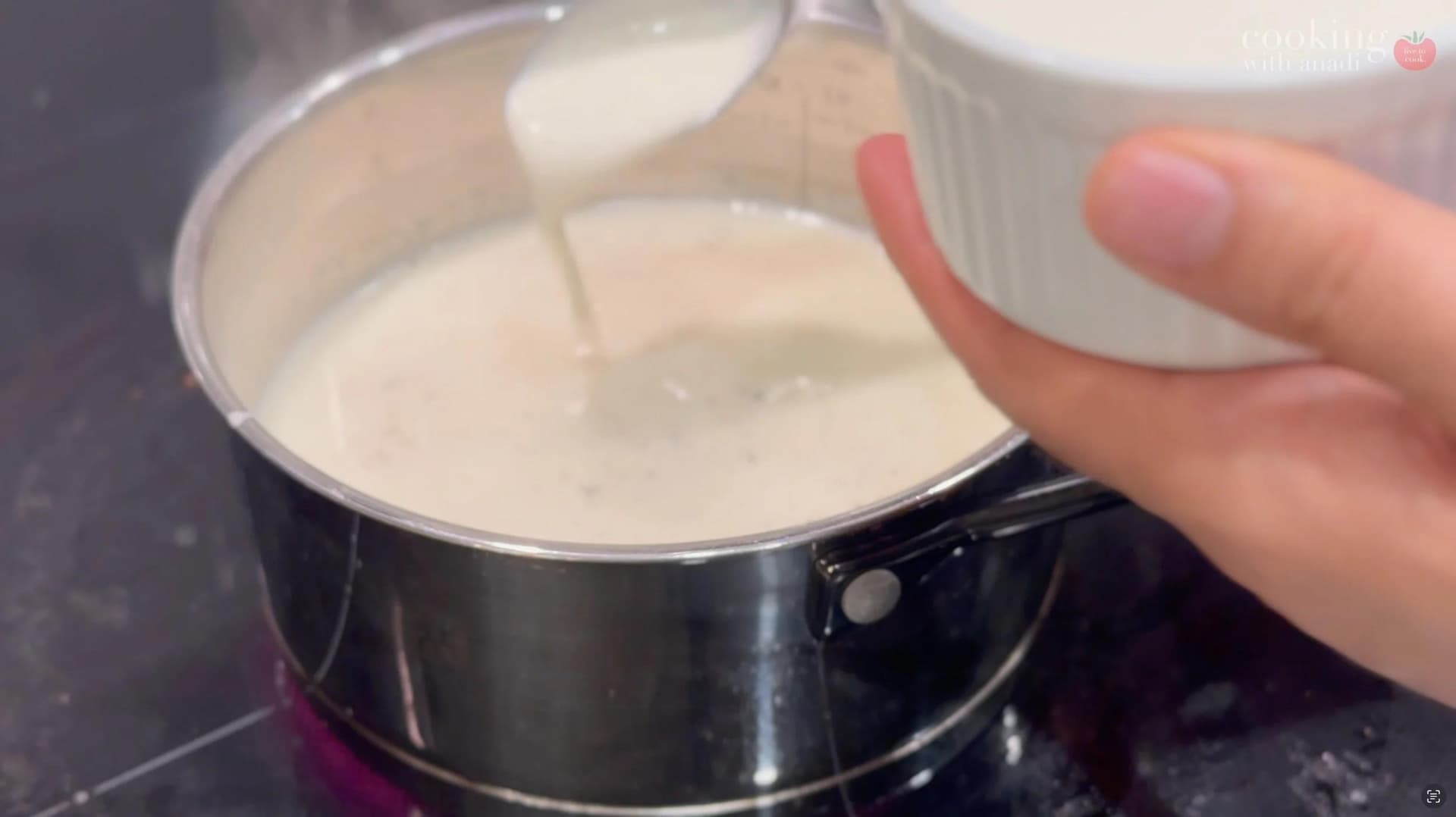 Milk being poured from a white pitcher into a black saucepan containing a cream-colored liquid mixture.