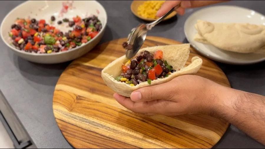 Hands filling a warm pita pocket with black bean and corn mixture topped with pico de gallo on a wooden board.
