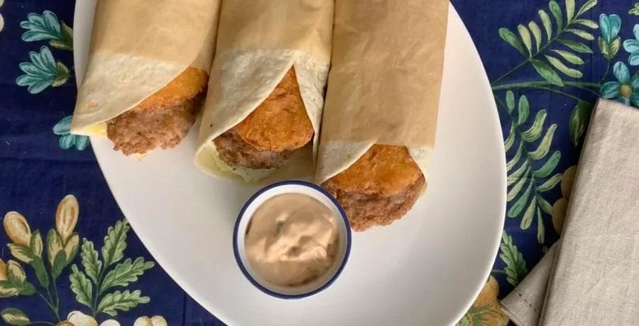 Three wrapped breakfast wraps with crispy edges on a white plate with a small cup of dipping sauce.