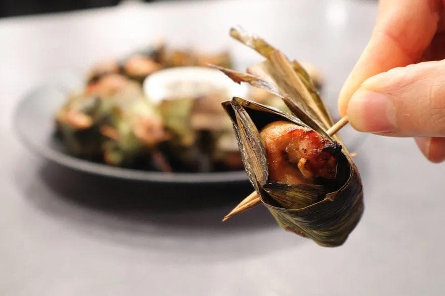 Hand holding a pandan leaf-wrapped chicken parcel secured with a toothpick, with more pieces on a dark plate in the background.