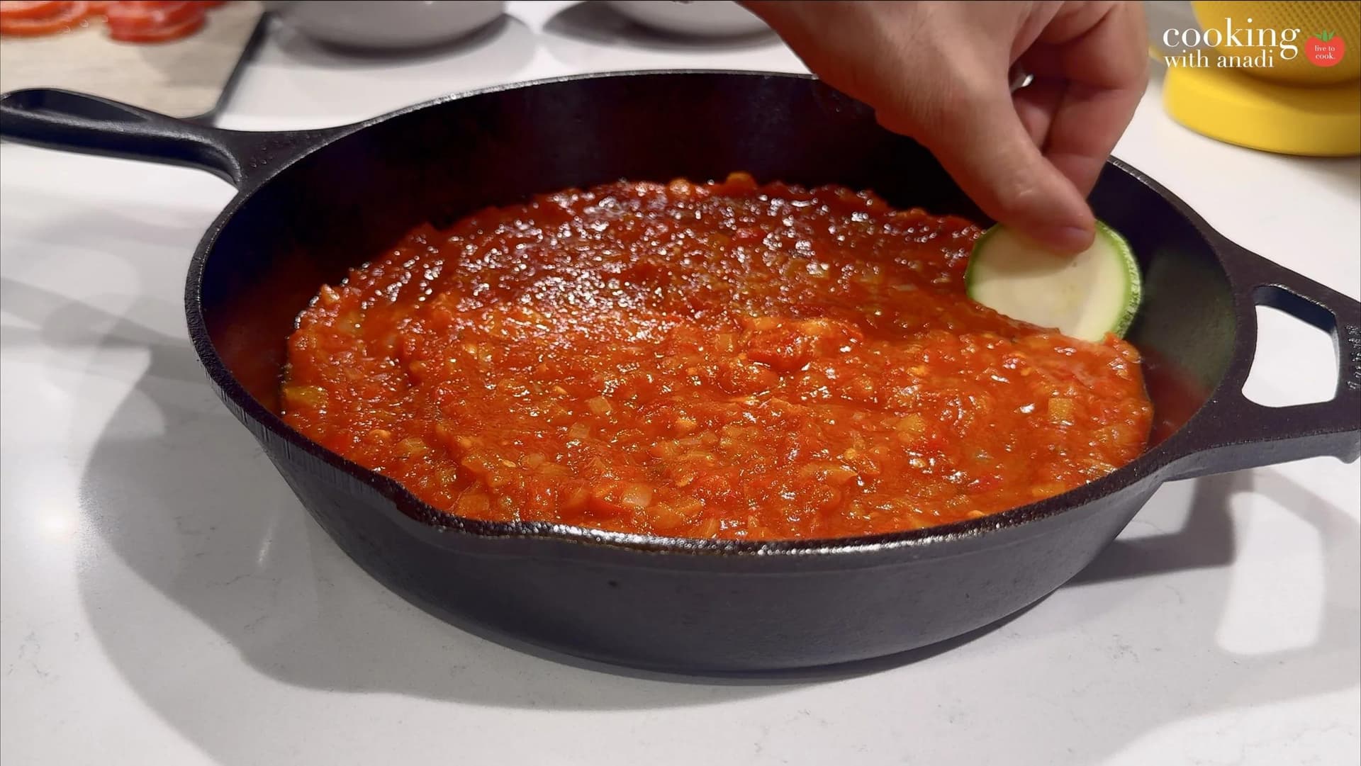 Cast iron skillet filled with chunky red piperade sauce, with a hand placing a zucchini round on top.