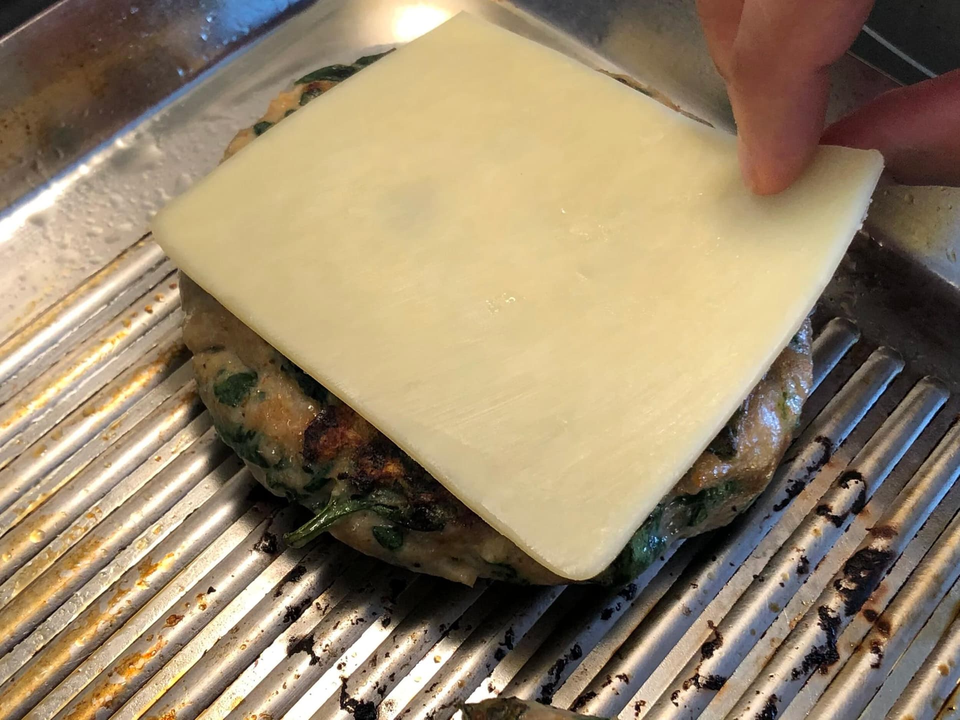 Melted cheese slice being placed on a cooked spinach turkey burger patty on a grill pan.