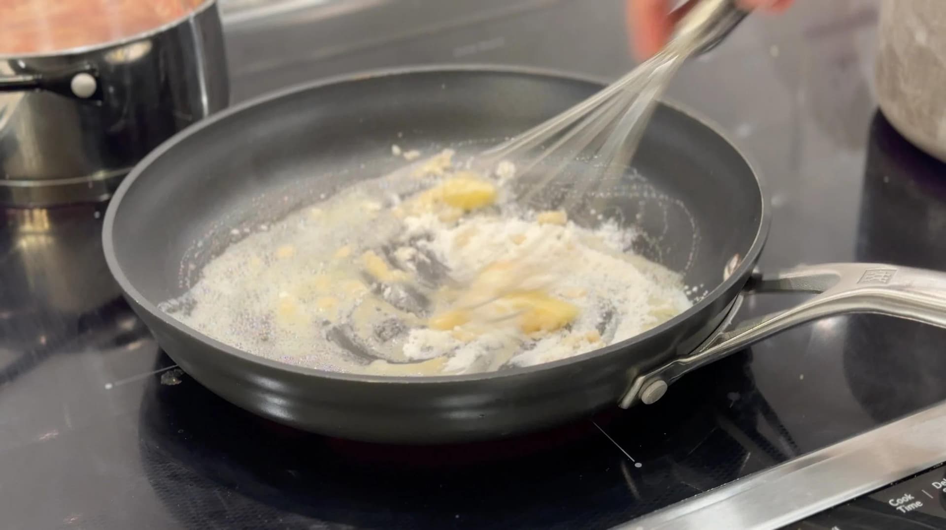 Whisking milk into a flour and butter roux in a skillet to make béchamel sauce for mac and cheese.